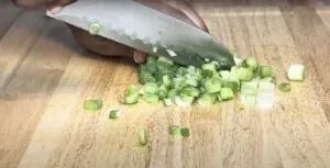 Person chopping green onions on a wooden cutting board with a knife.